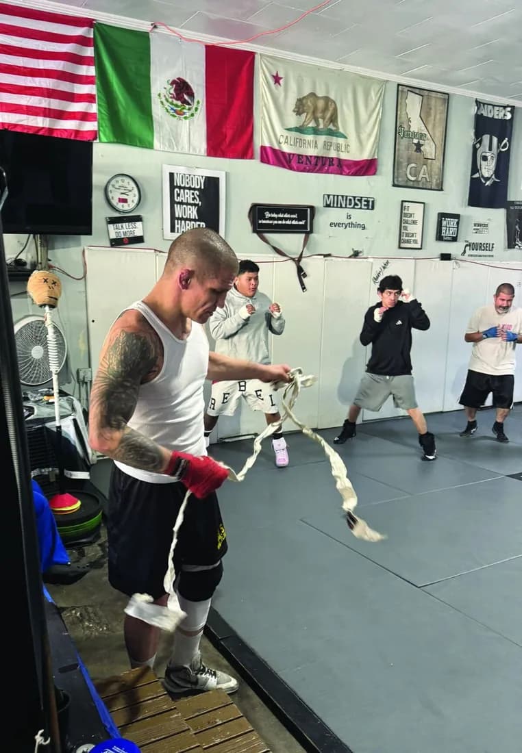 Bobby Taylor unrolling hand wraps on the gym floor while a class shadowboxes behind him